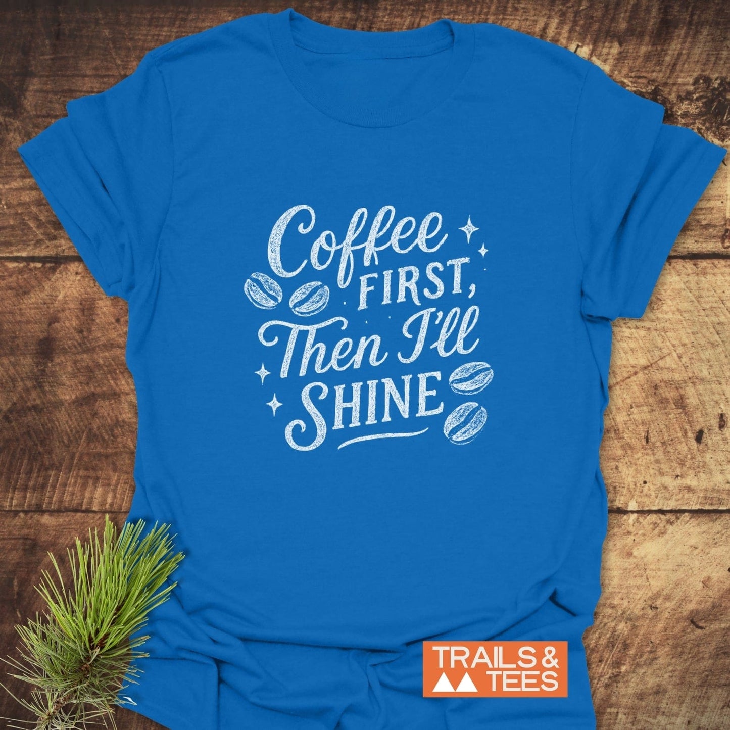 Coffee First T-Shirt featuring hand-lettered slogan with coffee-bean and sparkle illustrations, laid flat, showcasing ring-spun cotton designed for comfort and outdoor adventures.