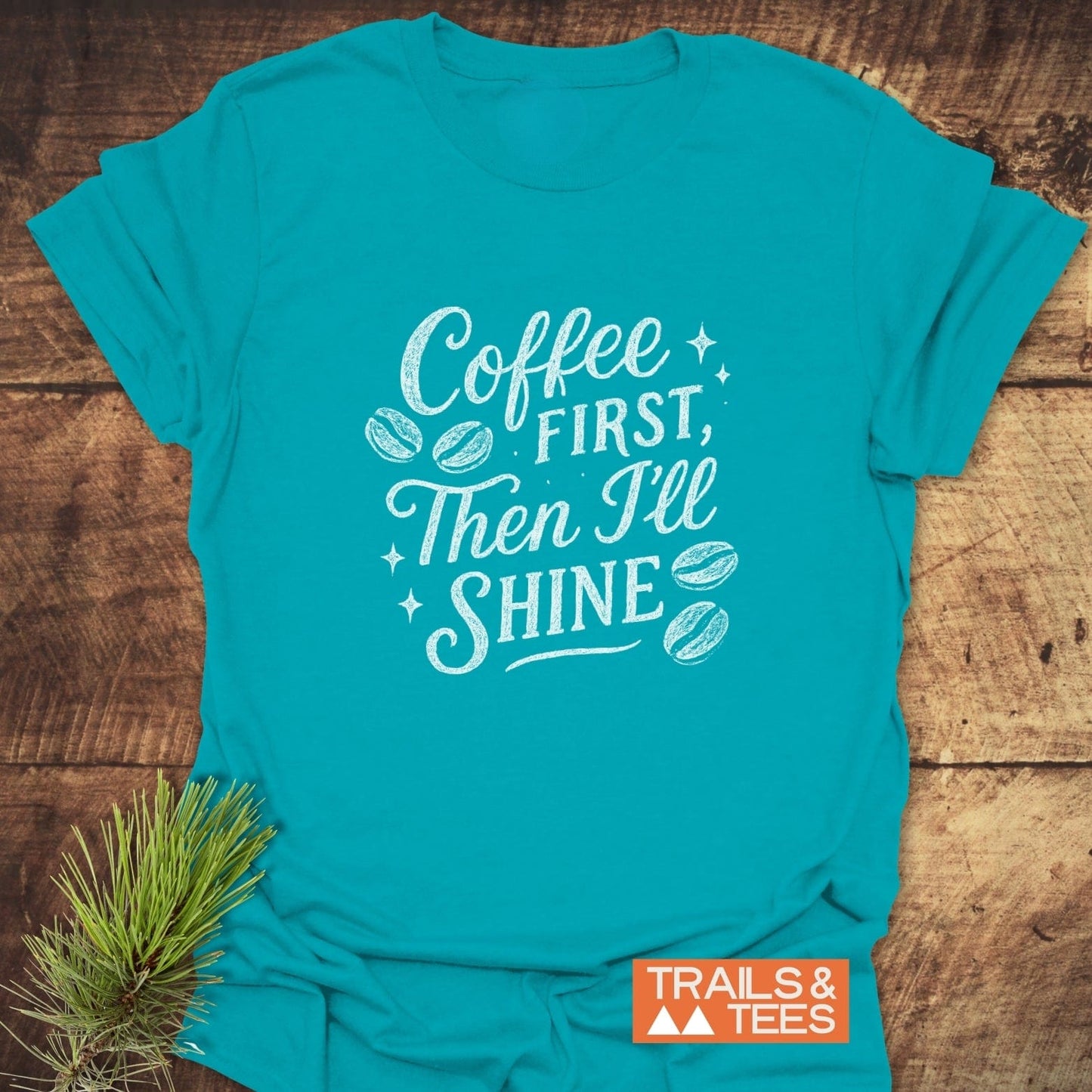 Teal Coffee First T-Shirt with slogan and graphics, featuring Trails & Tees logo. Pine sprig on wood surface. Made of soft, durable ring-spun cotton.