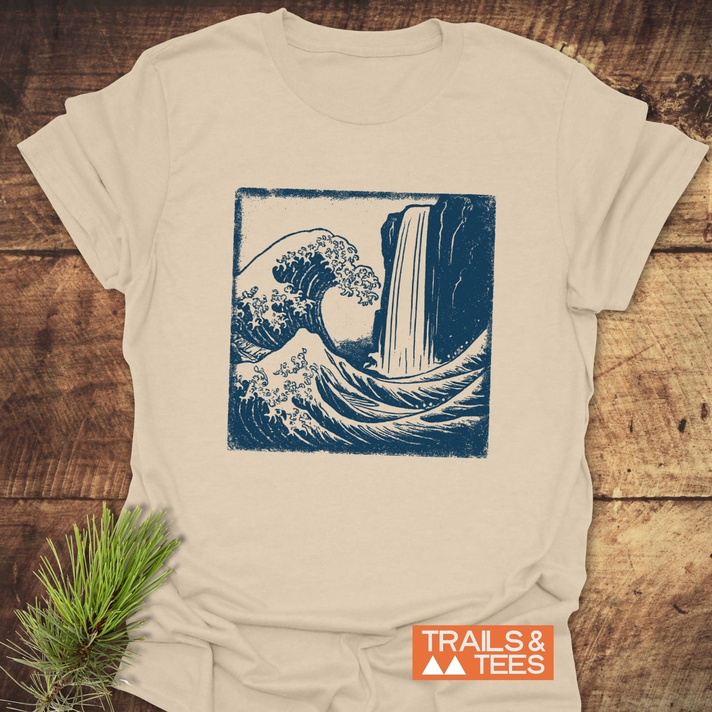 Waterfall And Wave T-Shirt on wood surface, featuring a deep blue ocean wave and waterfall graphic, pine sprig accent, and TRAILS & TEES logo nearby.