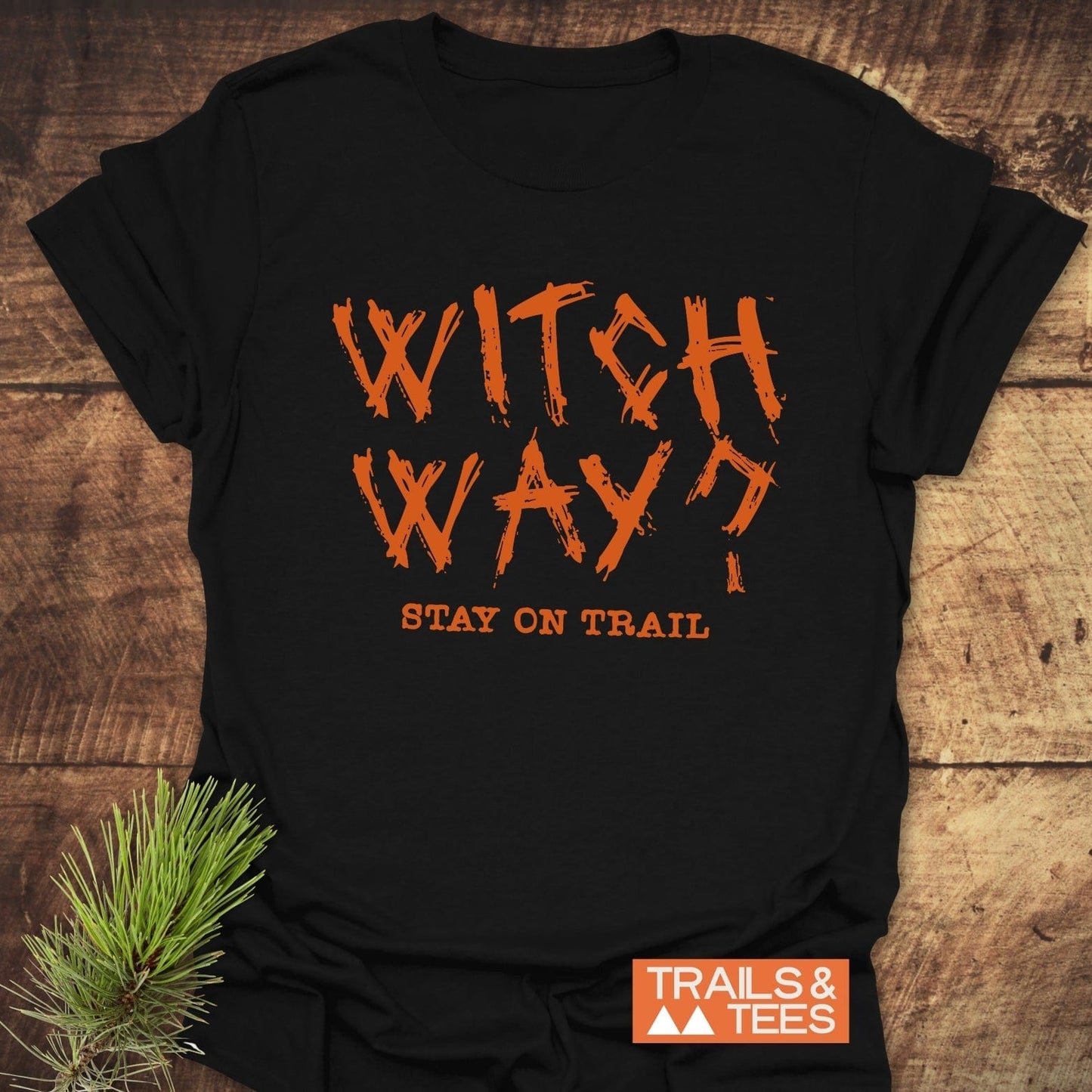 Witch Way? T-Shirt on wood surface, featuring bold orange text. Made from soft cotton, it offers durability with pre-shrunk fit and evergreen accent.