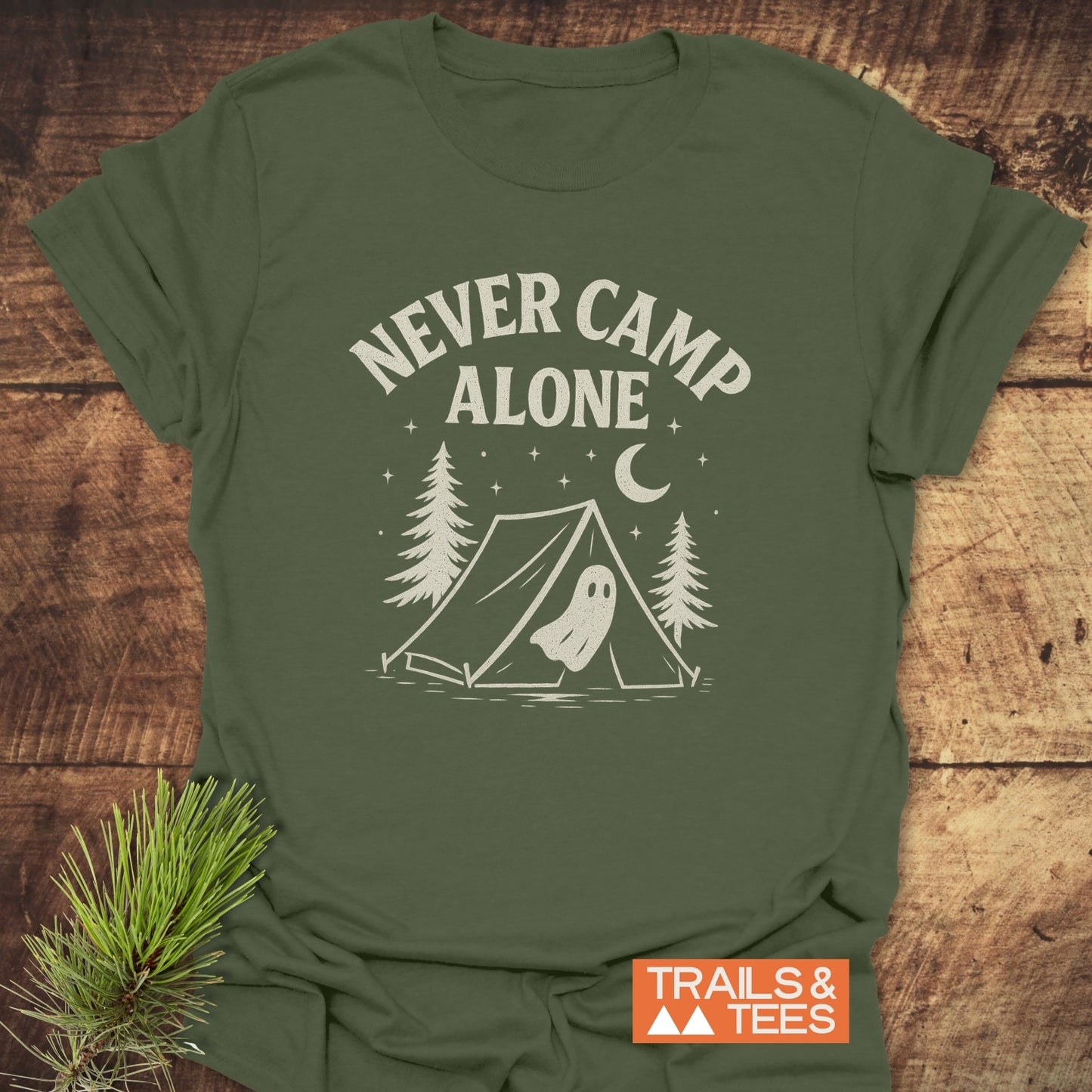 Never Camp Alone T-Shirt featuring a campsite graphic and slogan, laid flat with a pine sprig. Made from soft, durable cotton for outdoor adventures.