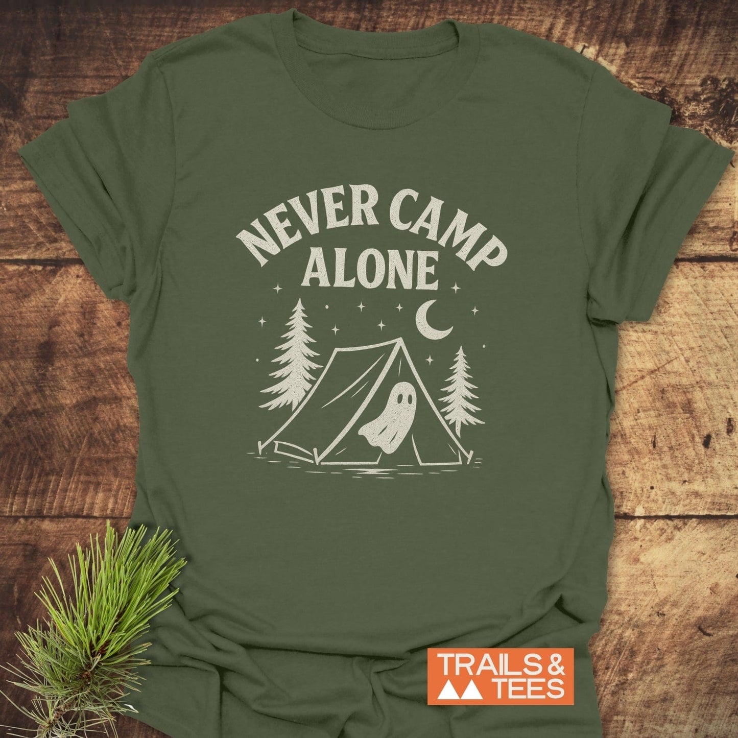 Never Camp Alone T-Shirt featuring a campsite graphic and slogan, laid flat with a pine sprig. Made from soft, durable cotton for outdoor adventures.