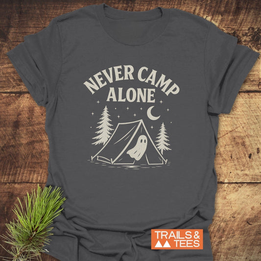 Never Camp Alone T-Shirt features a camp scene with trees, a tent, and a ghost under stars, crafted from soft cotton for outdoor enthusiasts.