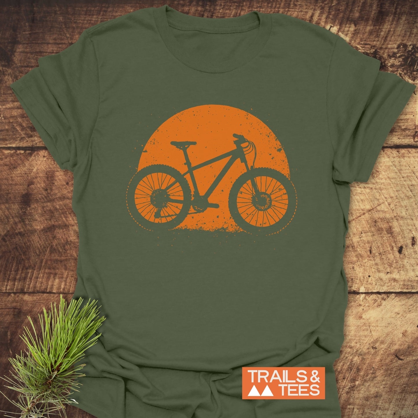 Grunge Mountain Bike T-Shirt featuring a mountain-bike silhouette and sun-like circle, laid on wood with pine sprig, embodying outdoor adventure and comfort.