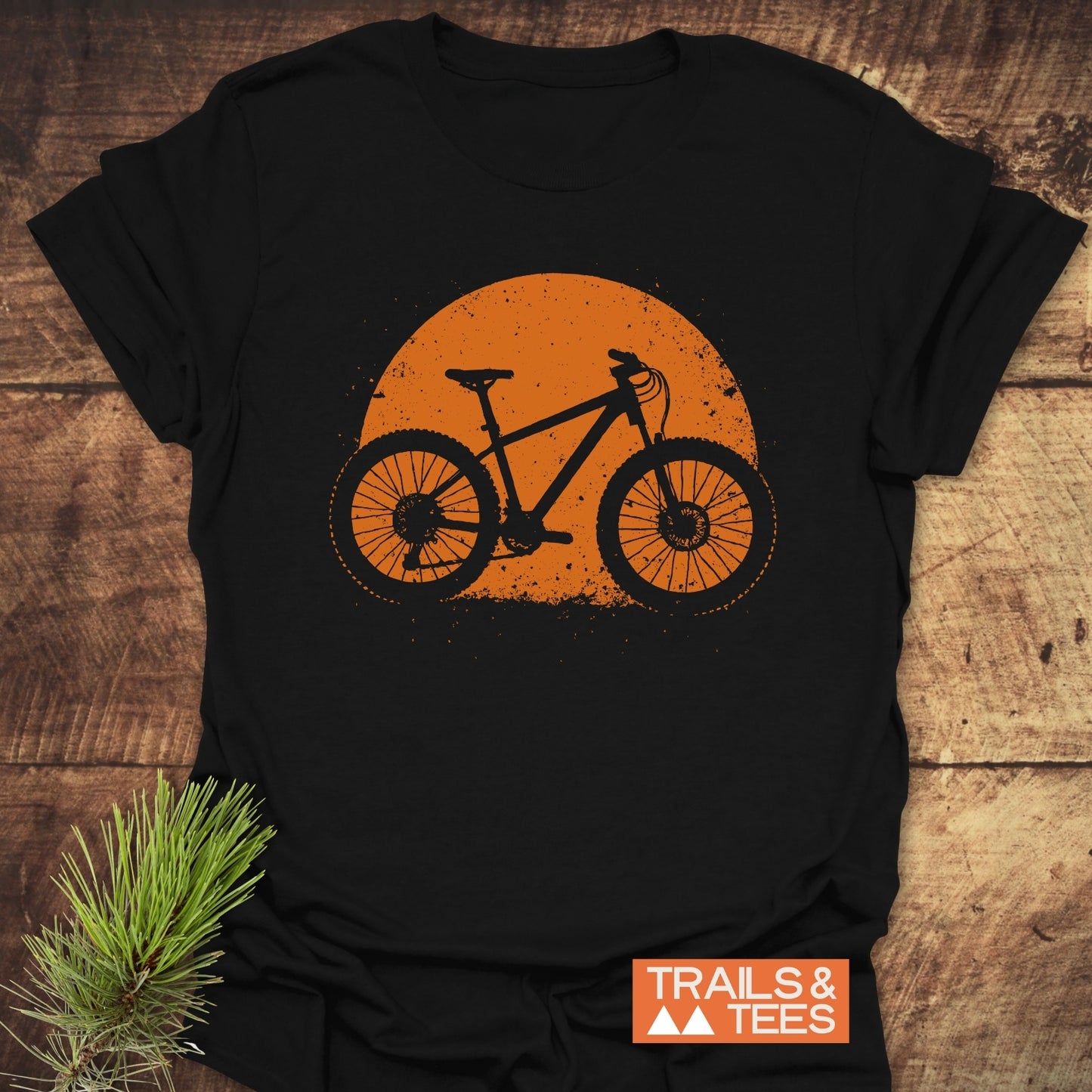 Grunge Mountain Bike T-Shirt featuring an orange bicycle graphic on black cotton, displayed on wood with pine accent, highlighting outdoor adventure style by TRAILS & TEES.
