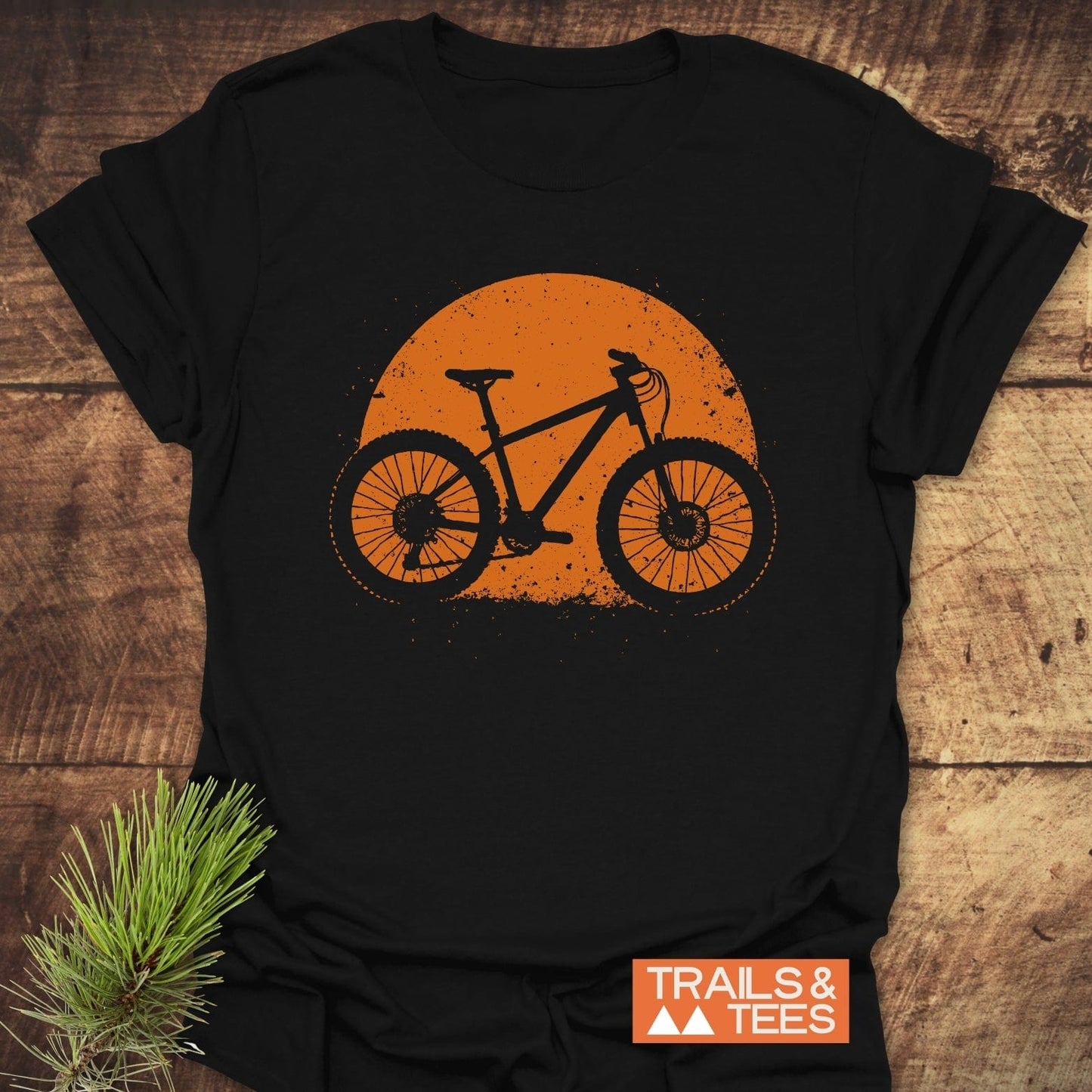 Grunge Mountain Bike T-Shirt featuring an orange bicycle graphic on black cotton, displayed on wood with pine accent, highlighting outdoor adventure style by TRAILS & TEES.
