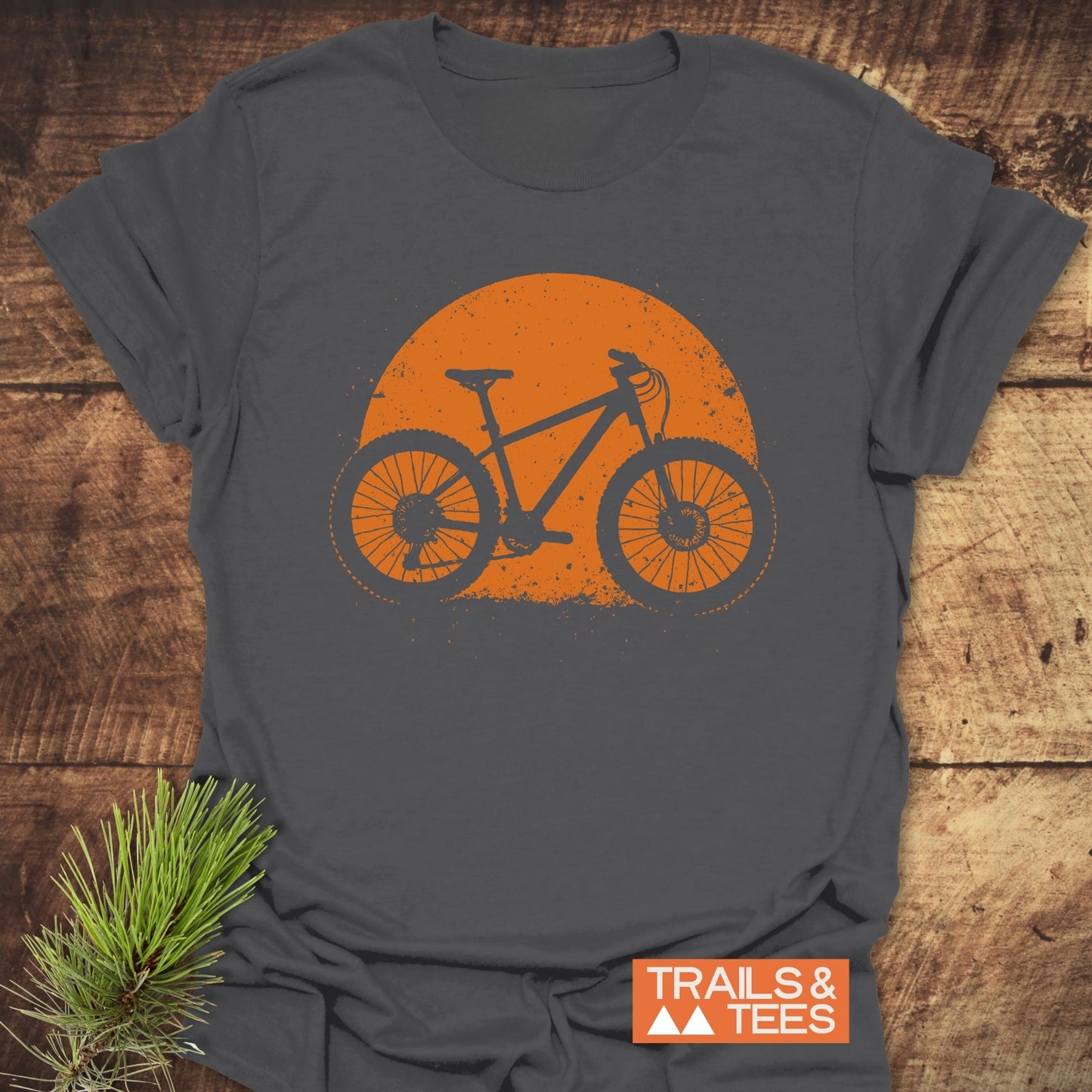 Grunge Mountain Bike T-Shirt featuring an orange bike silhouette on dark-gray fabric, styled with a pine sprig, embodying outdoor adventure spirit.