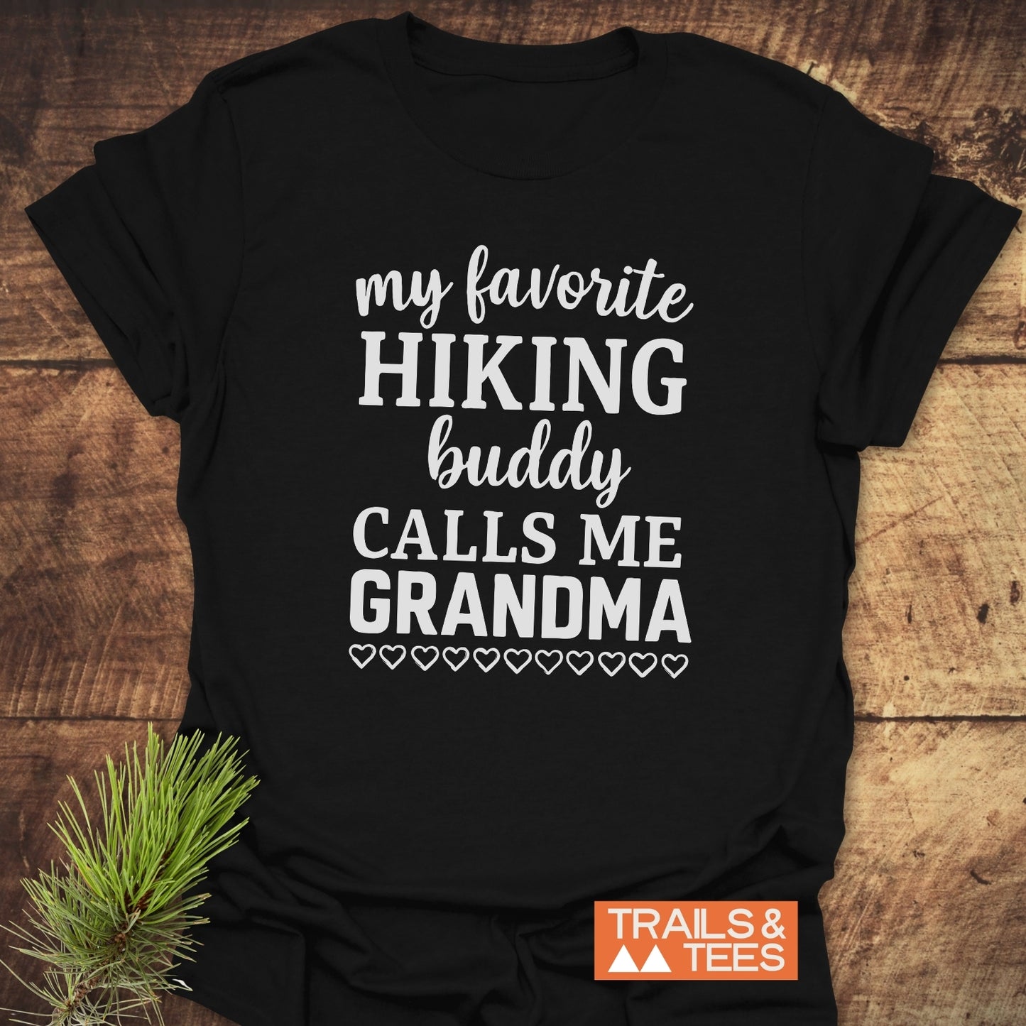 Black T-shirt with Hiking Grandma slogan and heart icons, on rustic wood. Features durable, lightweight cotton, perfect for outdoor adventures.