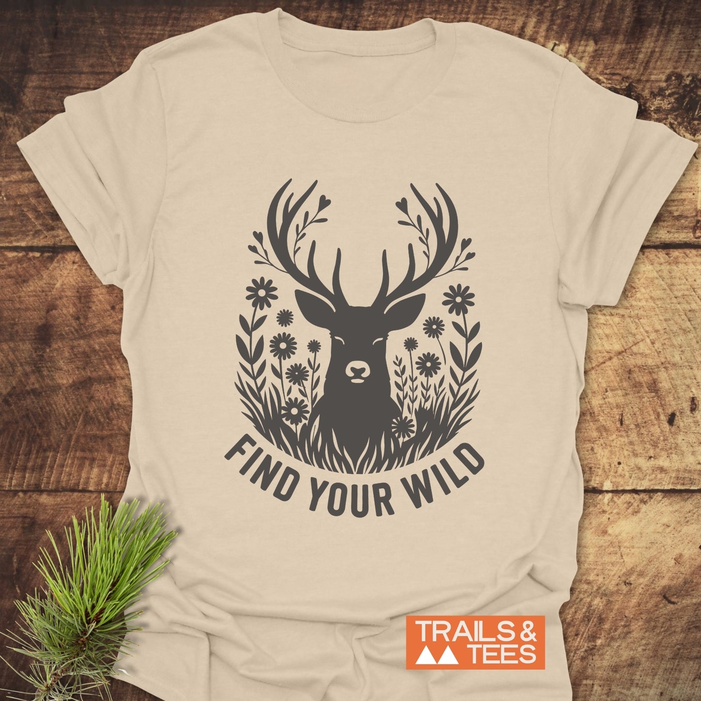 Find Your Wild T-Shirt featuring a deer design with antlers as leafy branches, wildflowers, and “FIND YOUR WILD” slogan. Includes Trails & Tees logo.