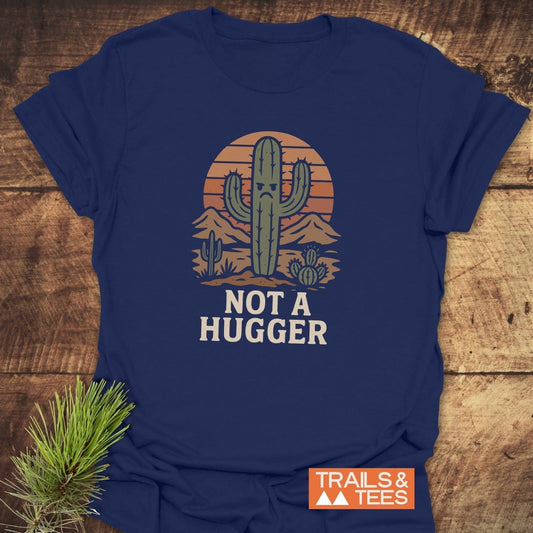 Navy-blue Not A Hugger T-shirt with a grumpy cactus desert scene, crafted from soft cotton, laid on a wooden surface, featuring TRAILS & TEES logo.