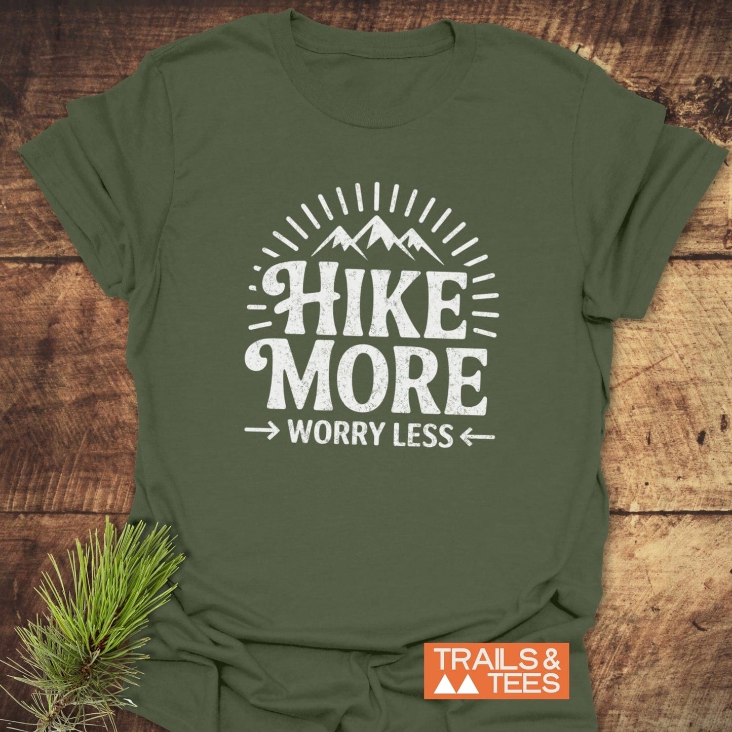 Hike More Worry Less T-Shirt featuring mountain graphic and slogan, laid on wood with pine sprig. Made from soft, pre-shrunk cotton for outdoor enthusiasts.