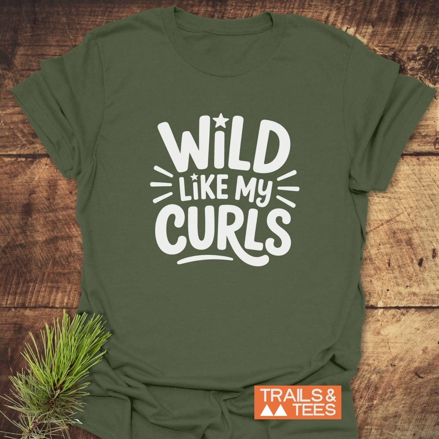 Wild Like My Curls T-Shirt in olive-green, featuring bold white lettering and nature motifs, laid flat with a pine sprig accent on rustic wood.