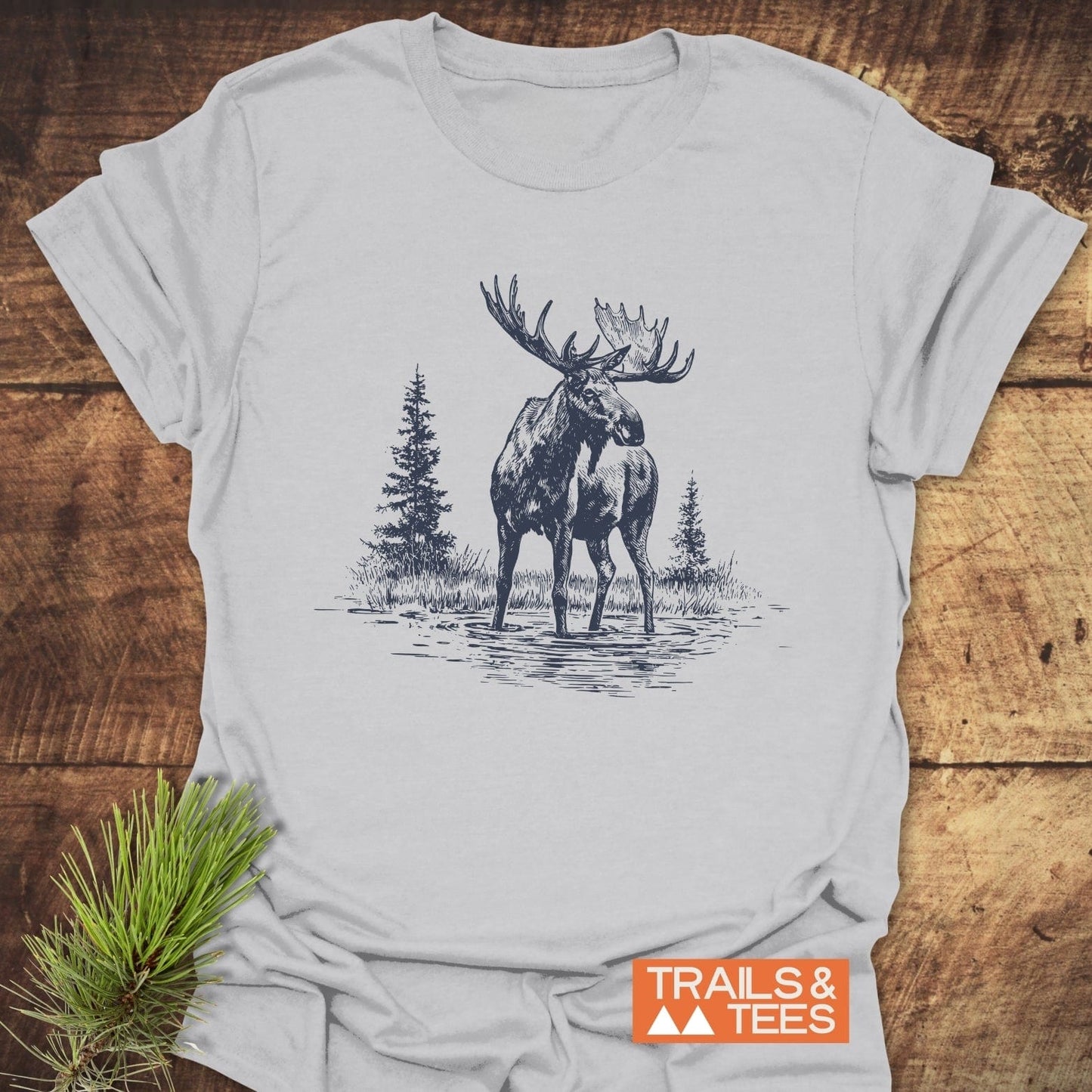 Moose T-Shirt with a sketch of a moose in water, evergreen trees behind. Laid on wood with a pine sprig, TRAILS & TEES logo visible.