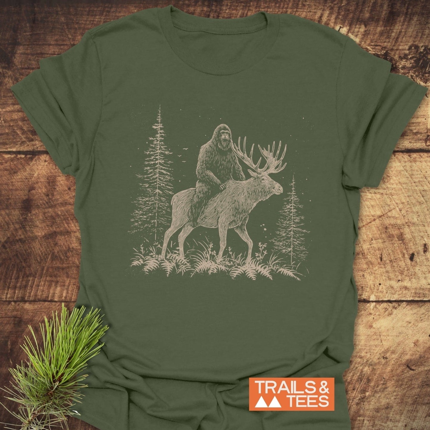 Best Friends Bigfoot T-Shirt displaying a cream sketch of Bigfoot on a moose, styled with pine sprig. Made from soft, pre-shrunk cotton for outdoor adventures.