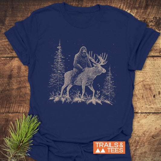 Best Friends Bigfoot T-Shirt featuring Bigfoot on a moose amid pines, printed on navy-blue cotton, offers soft comfort and durability with a “TRAILS & TEES” logo.