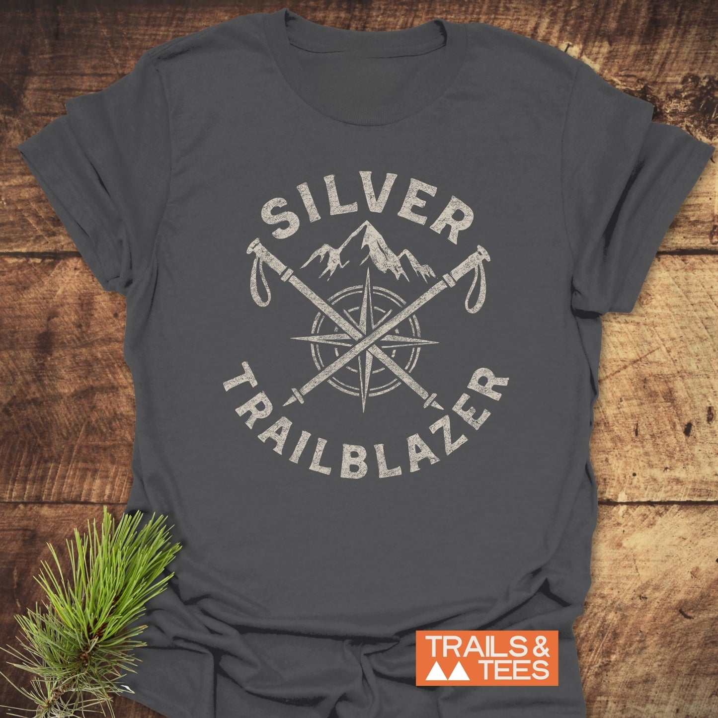Silver Trailblazer T-Shirt with distressed graphic featuring a mountain, compass, and crossed trekking poles, laid on wood. Includes TRAILS & TEES logo and pine sprig.