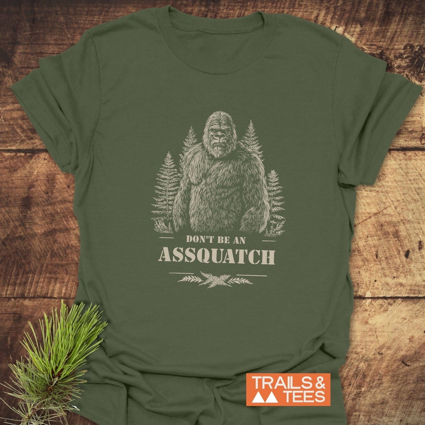 Assquatch T-Shirt featuring a stylized Sasquatch among pine trees with the slogan “DON’T BE AN ASSQUATCH,” crafted for comfort and adventure by Trails & Tees.