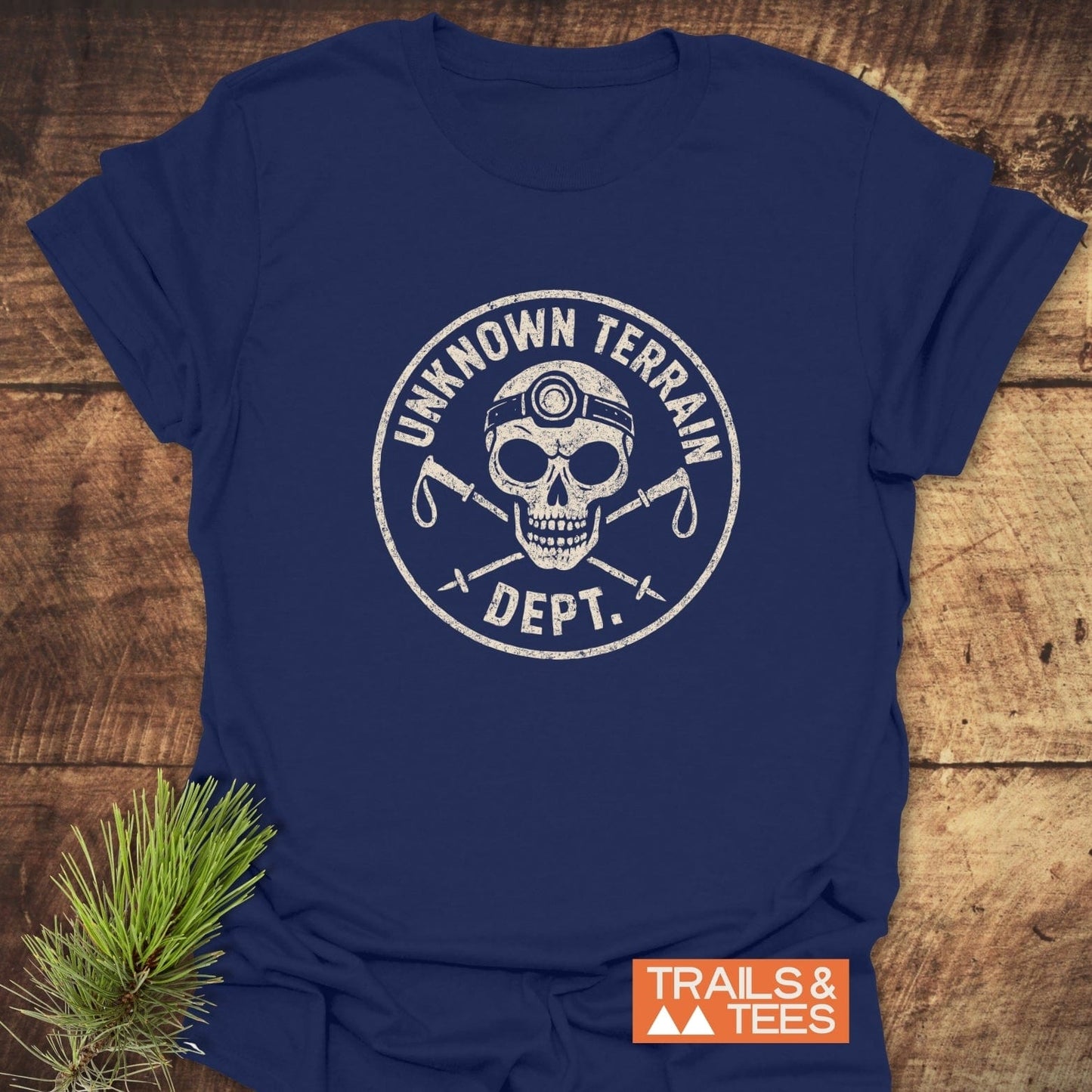 Unknown Terrain Dept. T-Shirt featuring a distressed skull logo with trekking poles, laid flat on wood, showcasing a durable, adventure-ready design.