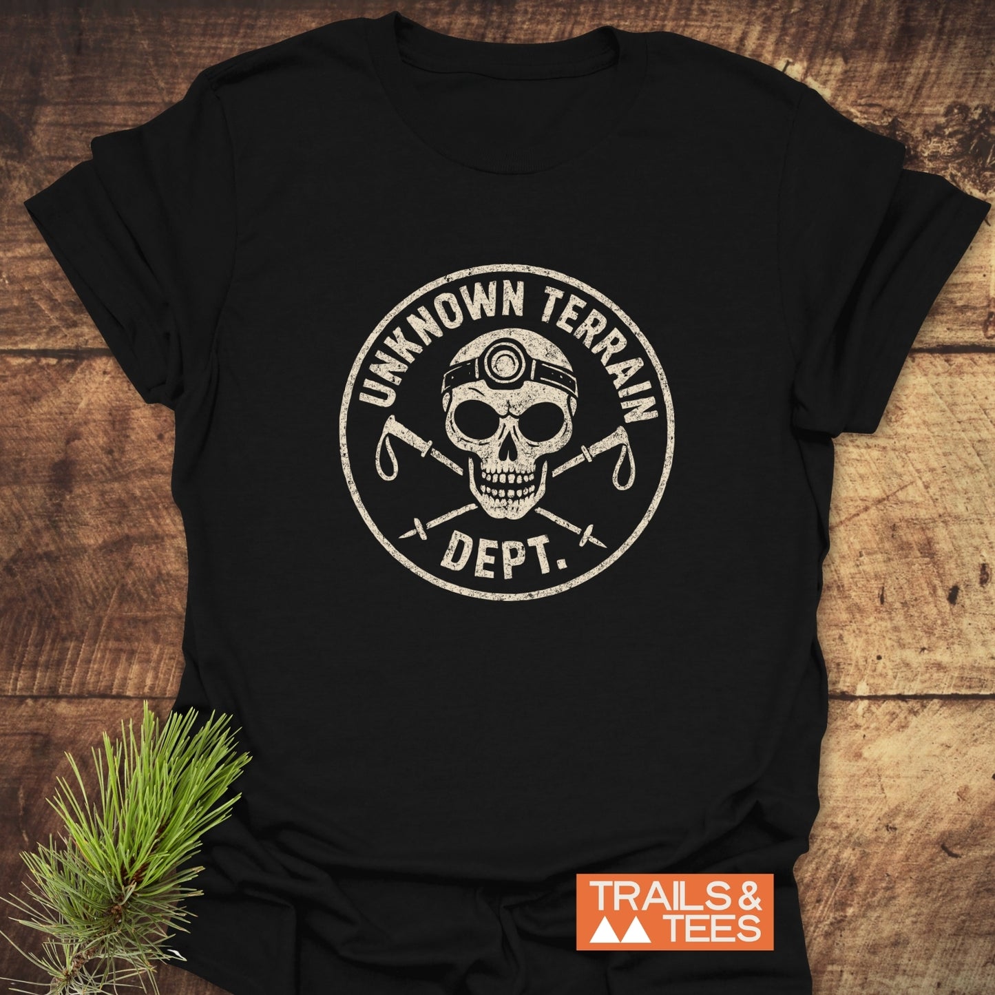 Unknown Terrain Dept. T-Shirt featuring a skull with a headlamp and crossed ice-axes, laid on wood. Includes “TRAILS & TEES” logo.