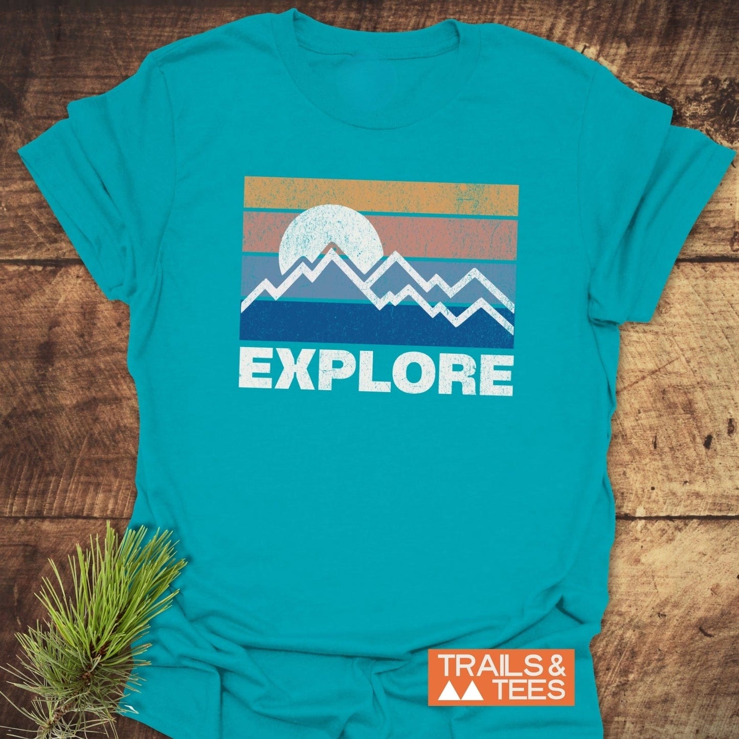Explore Retro Sunset T-Shirt featuring a graphic with mountains and EXPLORE text, laid on wood beside a pine sprig. Perfect for outdoor enthusiasts.
