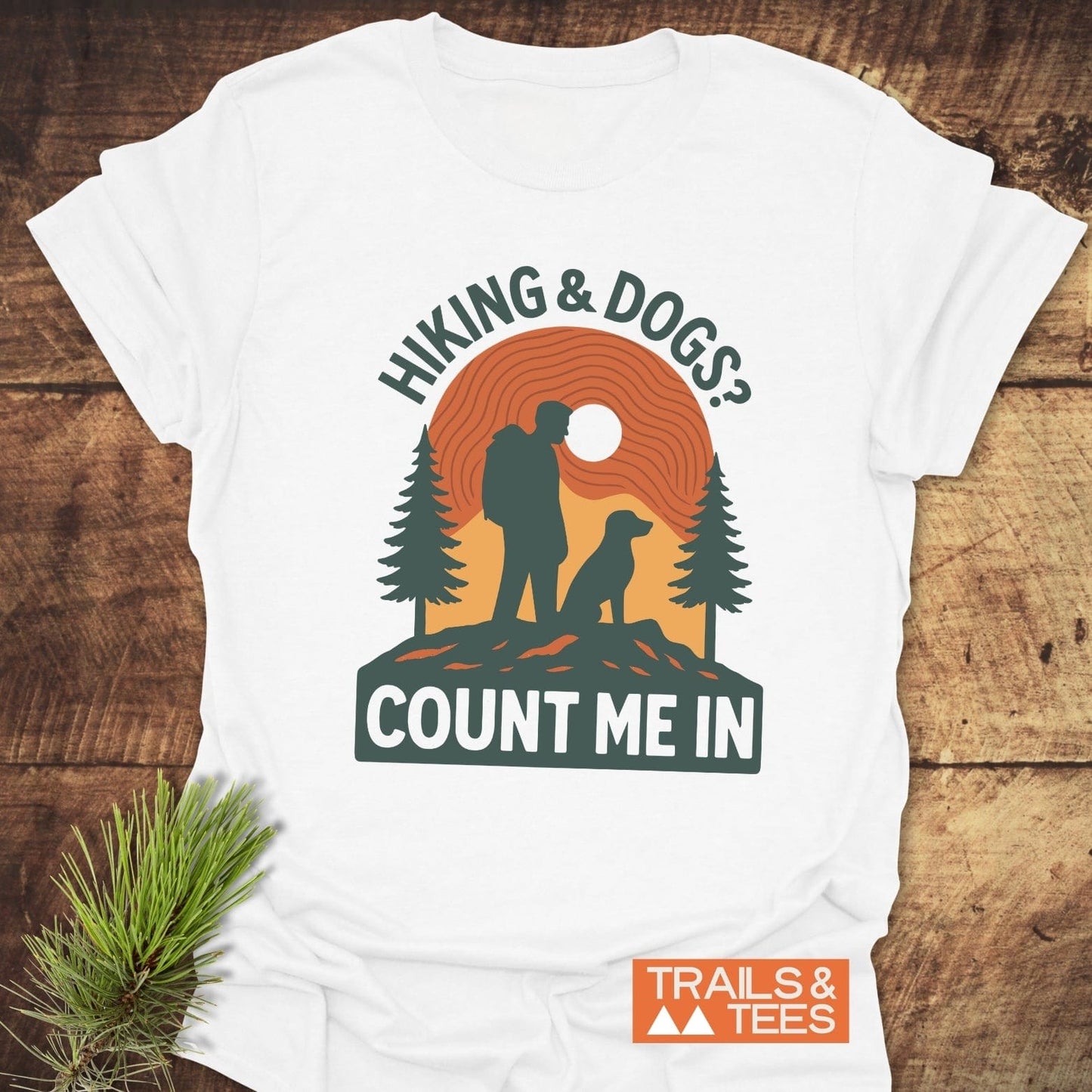 Hiking And Dogs T-Shirt featuring a hiker and dog design with pine trees and sun, laid on wood. Soft cotton, durable print, pre-shrunk fit.