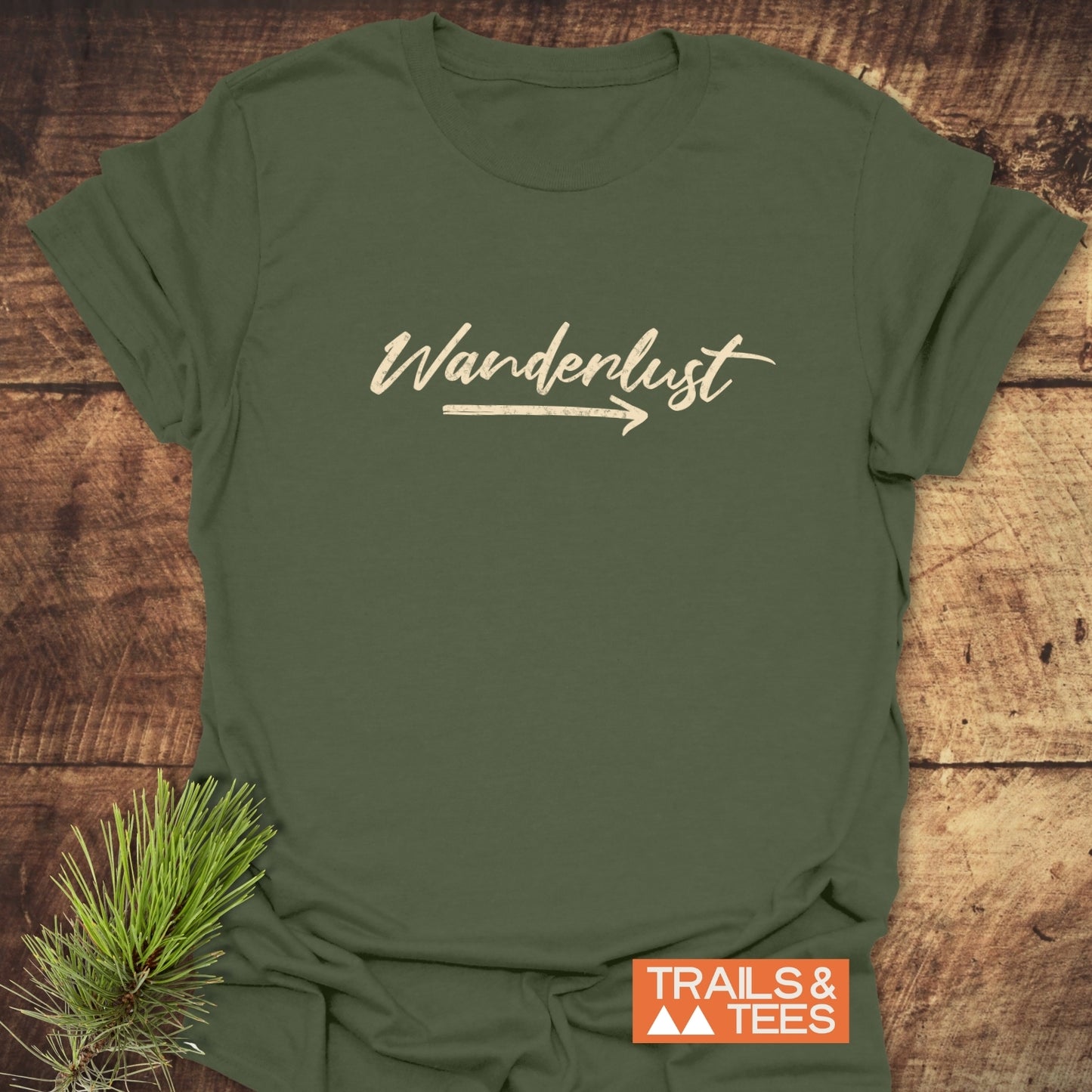 Wanderlust T-Shirt: Olive-green tee with Wanderlust script, arrow graphic, and pine sprig, symbolizing adventure. Includes TRAILS & TEES logo block.