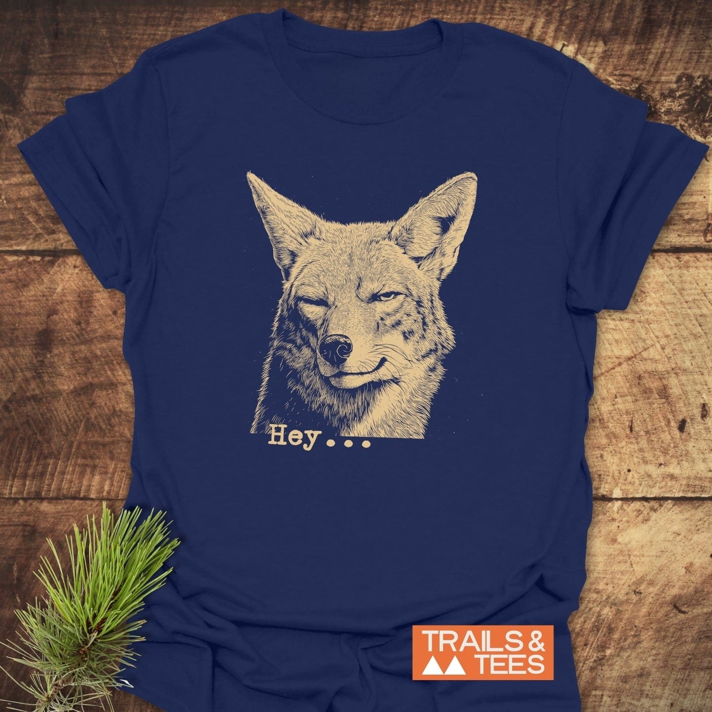 Hey... Coyote Funny T-Shirt with a winking canine illustration and Hey... in retro font. Laid flat on wood, highlighting its outdoor theme. Includes TRAILS & TEES logo and pine sprig accent.