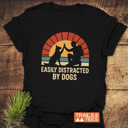 Easily Distracted By Dogs T-Shirt featuring a retro graphic of a person high-fiving a dog, sunset motif, and bold text on soft, ring-spun cotton.