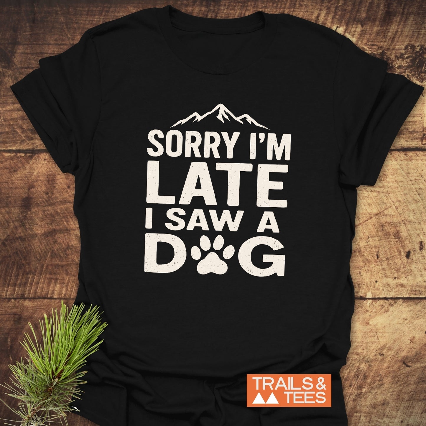 I Saw A Dog T-Shirt with a paw-print graphic and mountain silhouette, laid on wood. Features soft cotton, durable print, no side seams, by Trails & Tees.