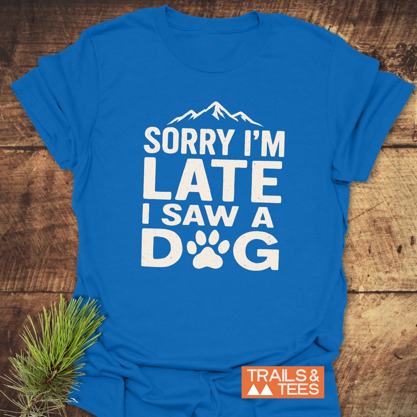 Bright blue I Saw A Dog T-shirt with mountain graphic and paw print, laid flat with a pine sprig beside it, featuring TRAILS & TEES label.
