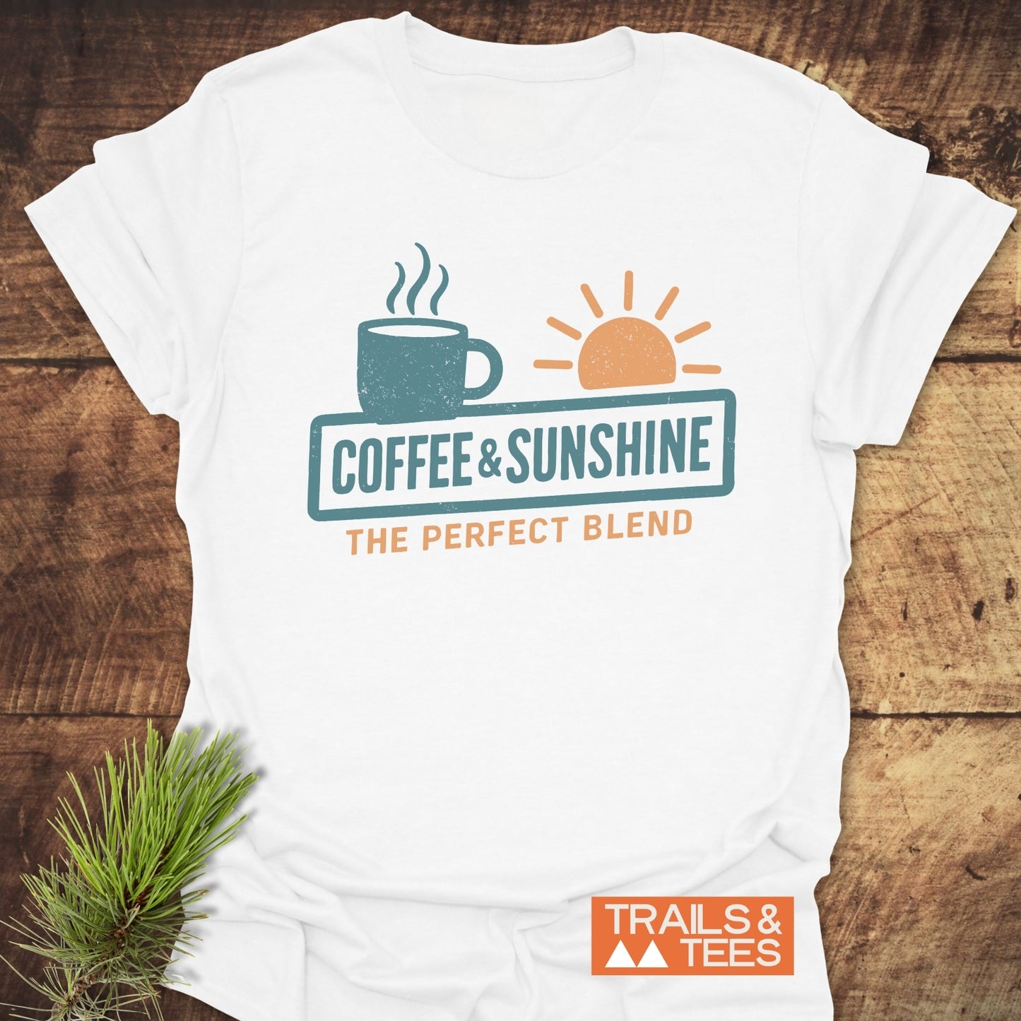 Coffee & Sunshine T-Shirt featuring a coffee mug and sun graphic, with The Perfect Blend slogan, lying on wood beside pine sprig and Trails & Tees logo.