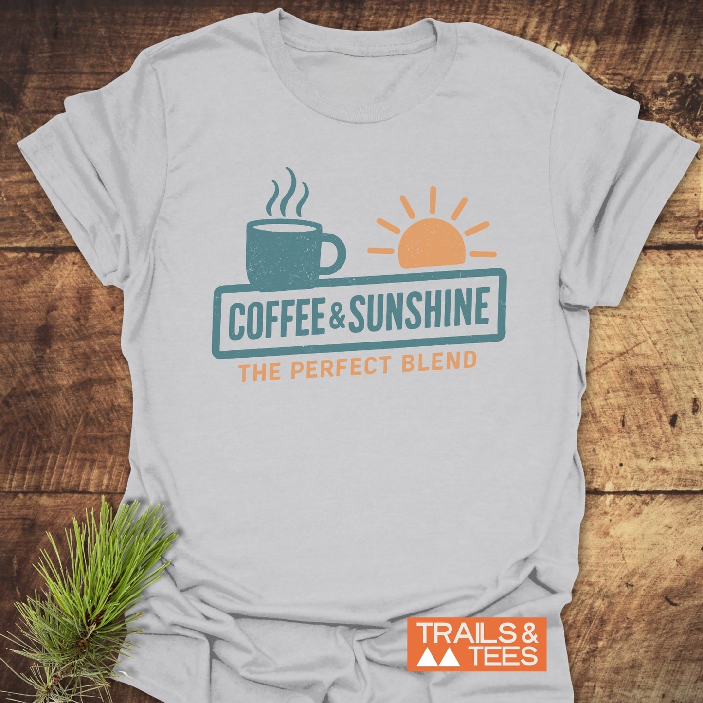 Coffee & Sunshine T-Shirt with stylized coffee mug and sun graphic, slogan, and Trails & Tees logo, displayed with an evergreen sprig on a wooden surface.