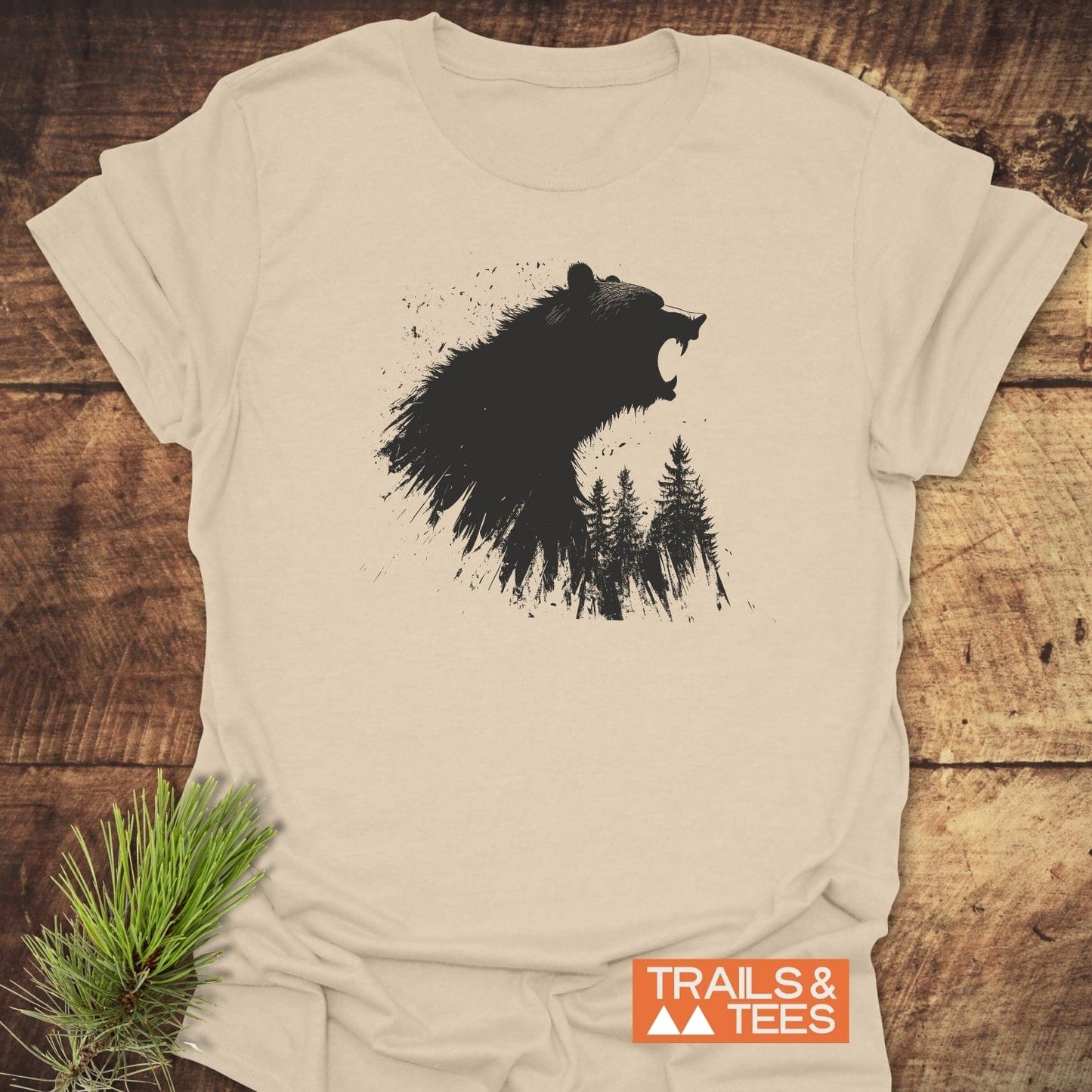 Light-beige Roaring Bear Forest T-Shirt featuring a black bear and pine trees graphic, laid on wooden surface with TRAILS & TEES logo beside it.