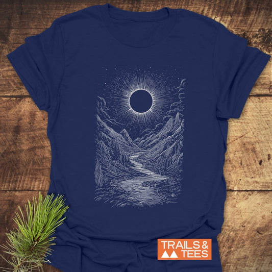 Eclipse Canyon T-Shirt with sun and mountain illustration, by Trails & Tees, on a wooden surface with pine bough, showcasing adventure-inspired design and comfort.