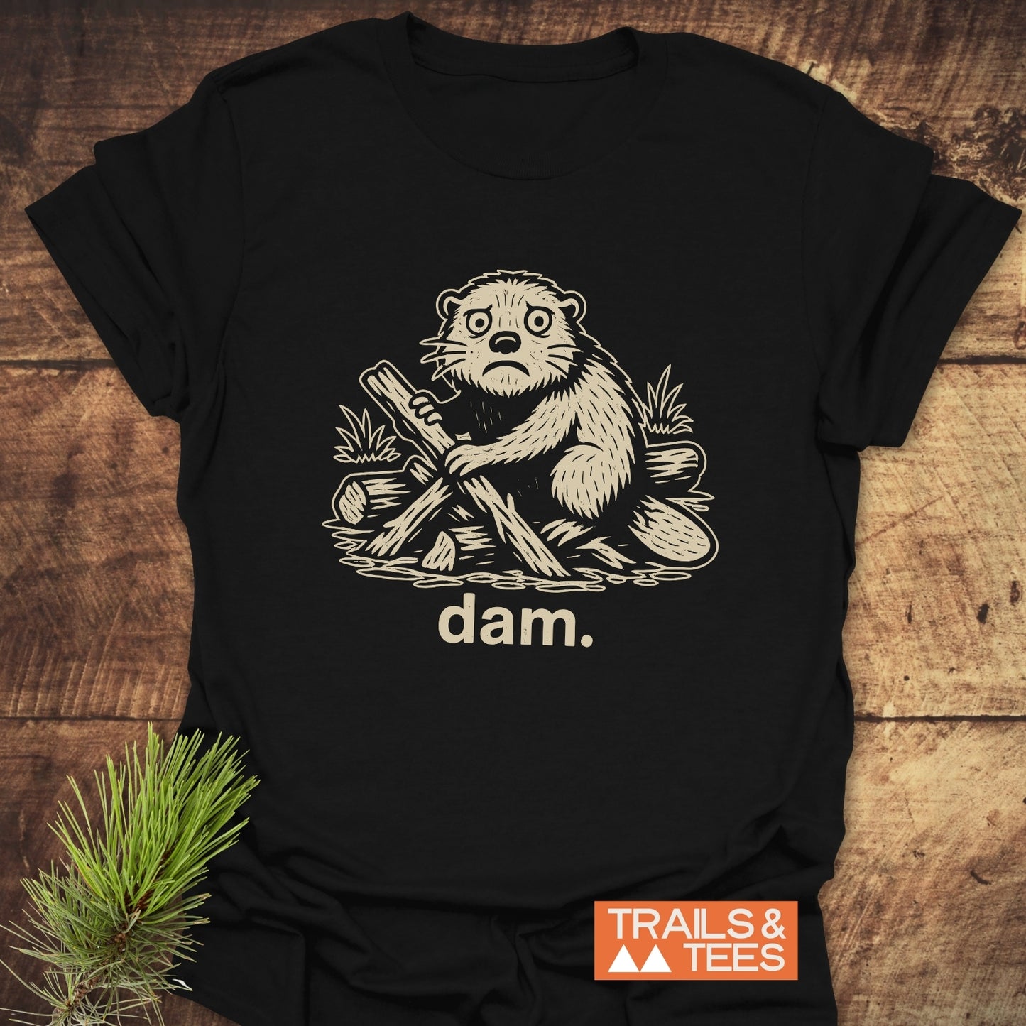 Beaver - Dam Funny T-Shirt featuring a cream beaver drawing, dam. text, and Trails & Tees logo on black cotton, displayed with a pine branch.