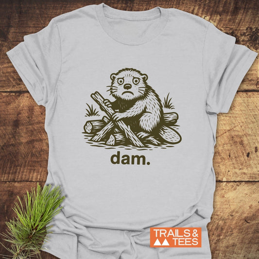 Light-gray T-shirt featuring a forlorn cartoon beaver with logs, “dam.” text, and “TRAILS & TEES” logo, highlighting comfort and outdoor adventure appeal.