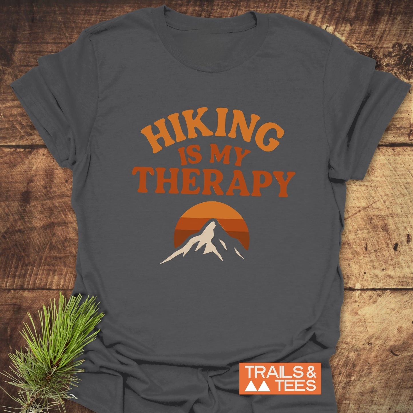 Hiking Therapy T-Shirt with HIKING IS MY THERAPY and mountain graphic, made of soft ring-spun cotton, laid flat on wood surface with pine sprig accent.