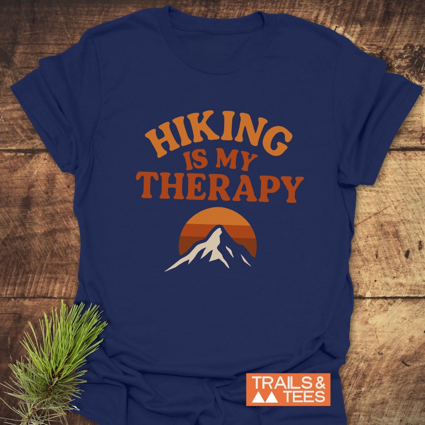 Navy-blue Hiking Therapy T-Shirt with a mountain sunset graphic and slogan, laid on wood. Crafted from soft cotton, it offers comfort and durability.