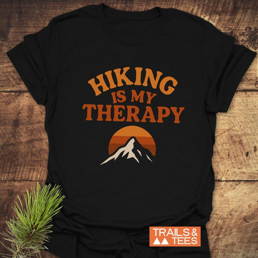 Hiking Therapy T-Shirt featuring HIKING IS MY THERAPY slogan and mountain graphic, laid on a wooden surface with pine sprig, showcasing outdoorsy appeal.