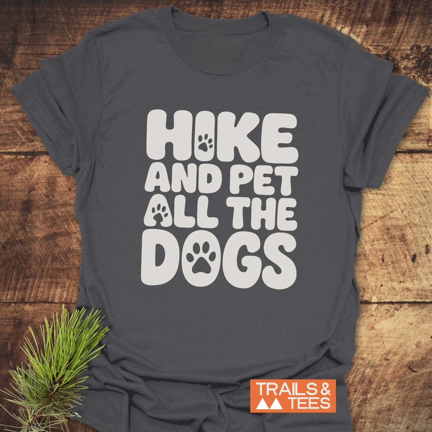 Charcoal-gray Hike And Pet Dogs T-shirt with paw-print letters, laid on wood with pine sprig. Soft ring-spun cotton, durable print, pre-shrunk for fit.