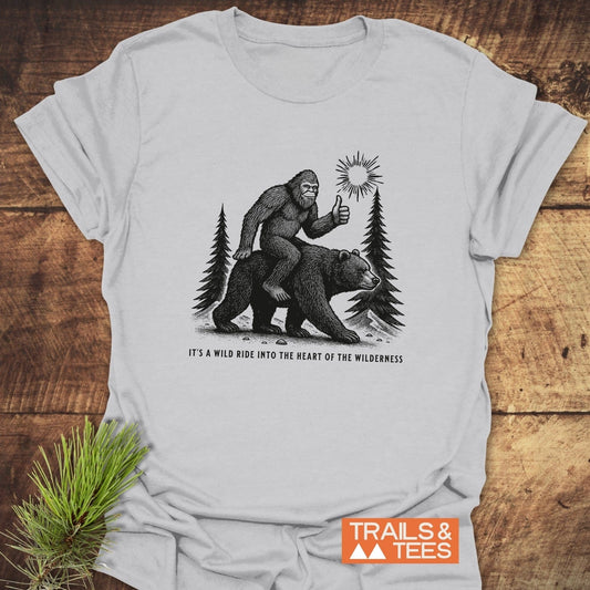 Wild Ride Bigfoot T-Shirt: Light-gray tee featuring Bigfoot on a bear under pine trees, with “IT’S A WILD RIDE INTO THE HEART OF THE WILDERNESS” slogan.