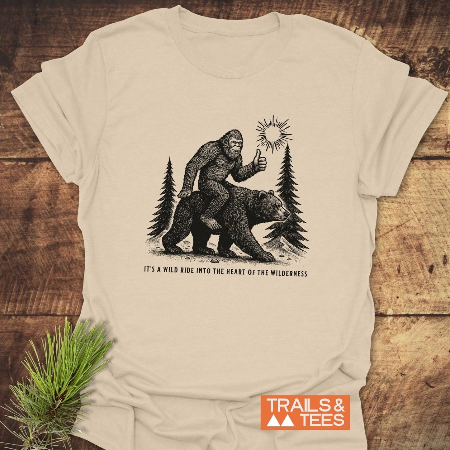 Wild Ride Bigfoot T-Shirt featuring Sasquatch on a bear through forest scene, slogan beneath. Made from soft, durable cotton for outdoor adventures.
