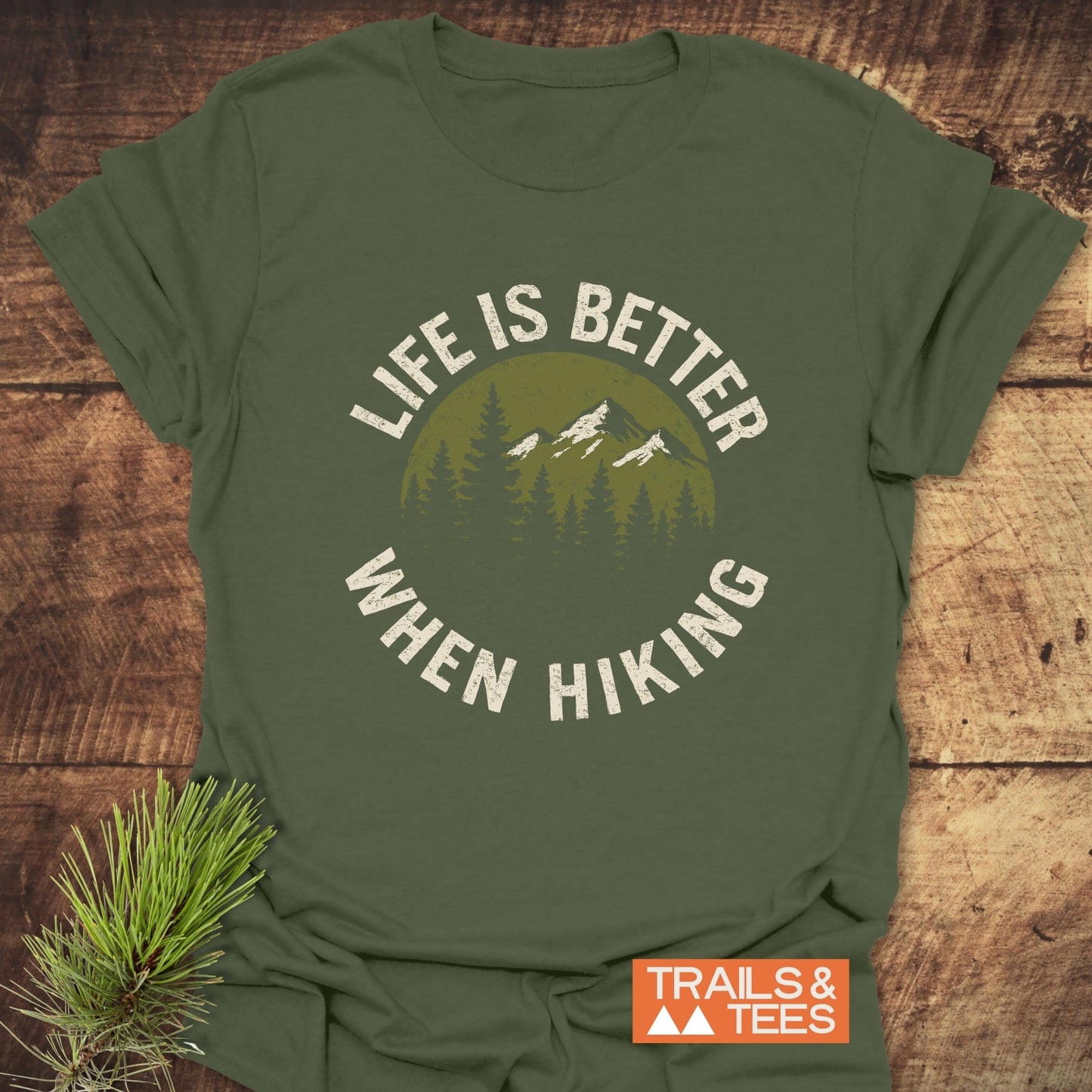 Life Is Better When Hiking T-Shirt with mountain and forest graphic, laid flat on a wooden surface, highlighting its outdoor adventure theme.
