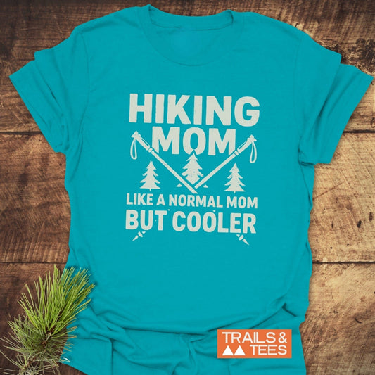 Teal-blue Hiking Mom T-Shirt with white graphic of crossed trekking poles and pine trees. Features slogan: Hiking Mom, Like a normal mom but cooler.