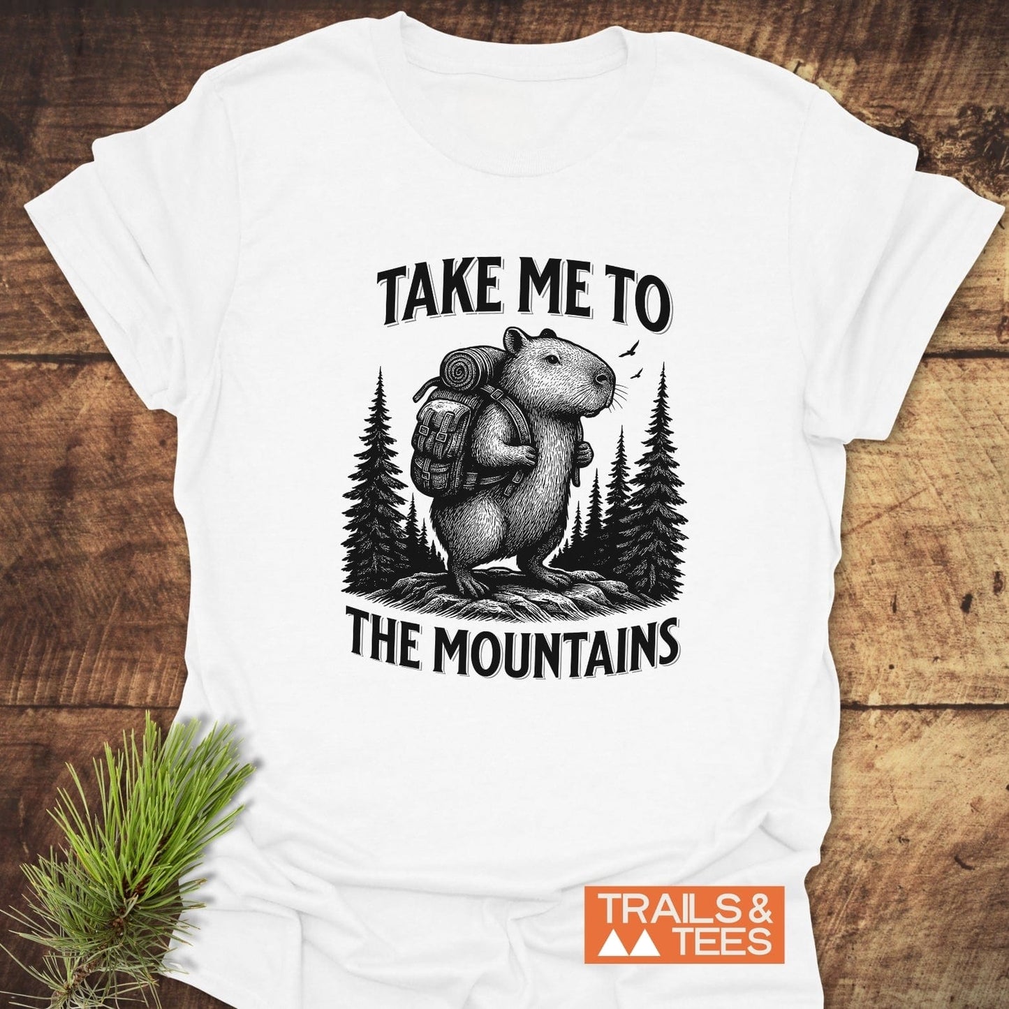 Capybara Mountains T-Shirt featuring a rodent with a hiking backpack amid trees. Soft, ring-spun cotton for comfort, pre-shrunk, and durable print.
