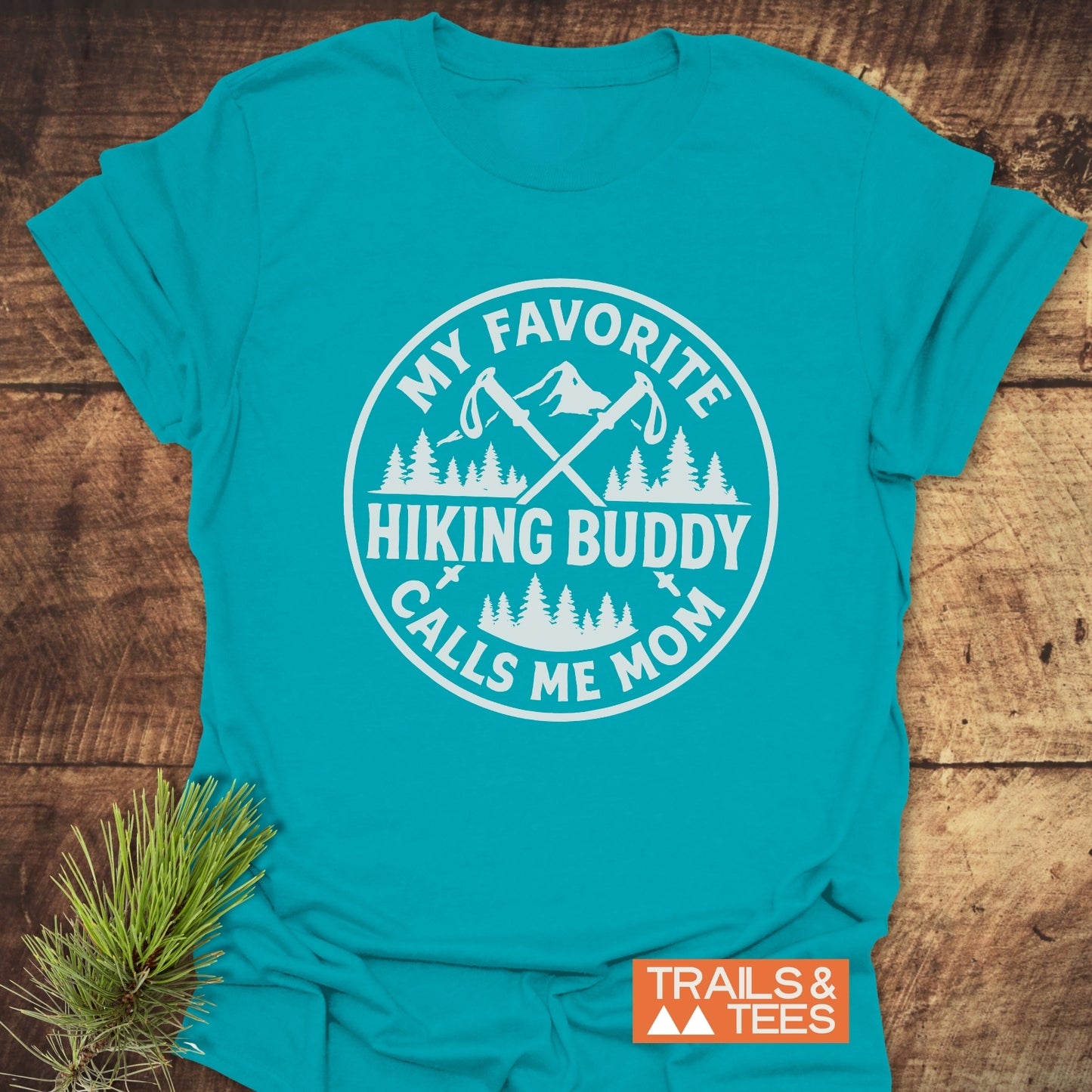 Teal T-shirt with hiking graphic and My Favorite Hiking Buddy Calls Me Mom slogan, showcasing Trails & Tees logo beside a pine sprig.