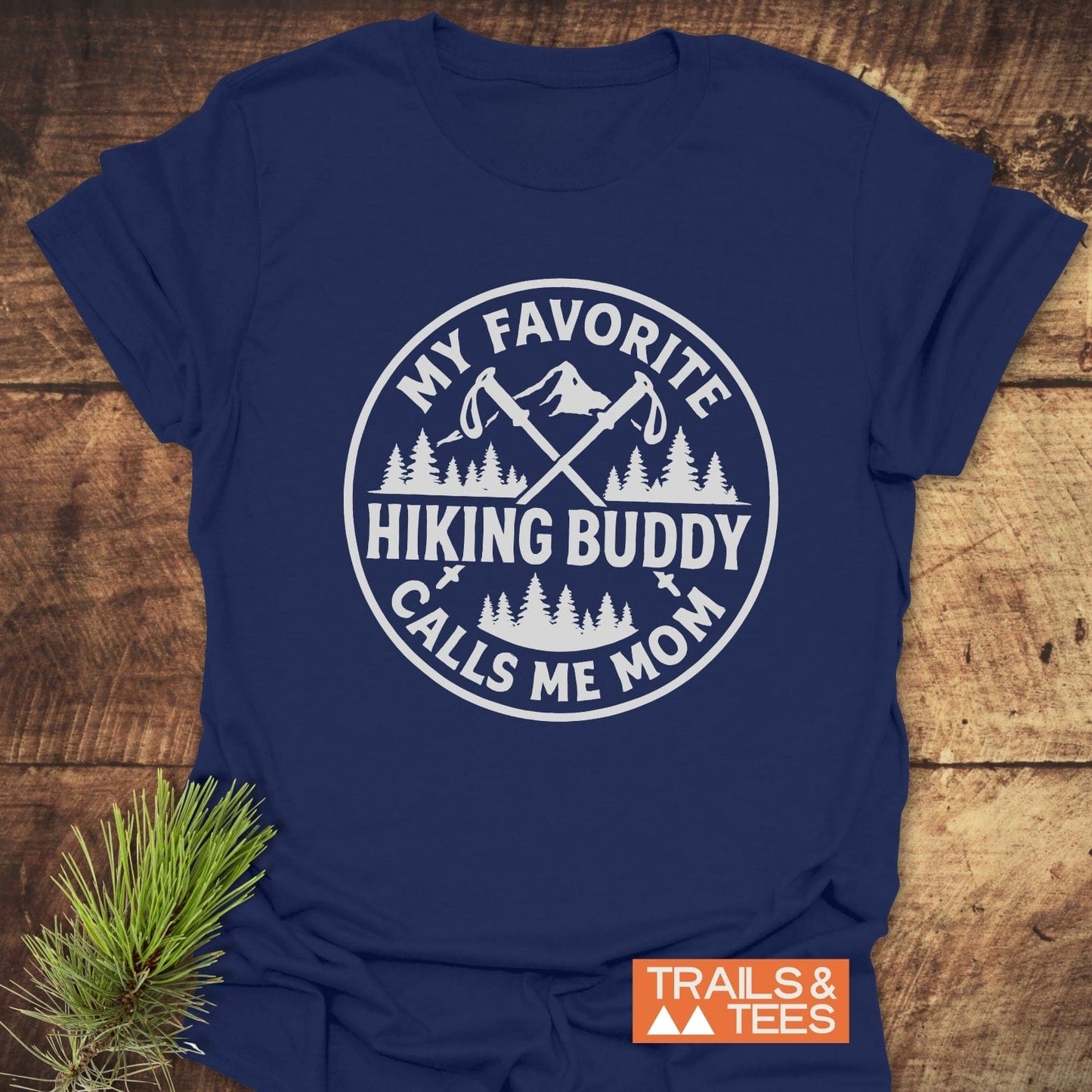 Navy-blue Hiking Buddy Mom T-Shirt featuring a mountain-themed emblem with trekking poles, laid on rustic wood. Designed for outdoor enthusiasts, crafted from soft, durable cotton.