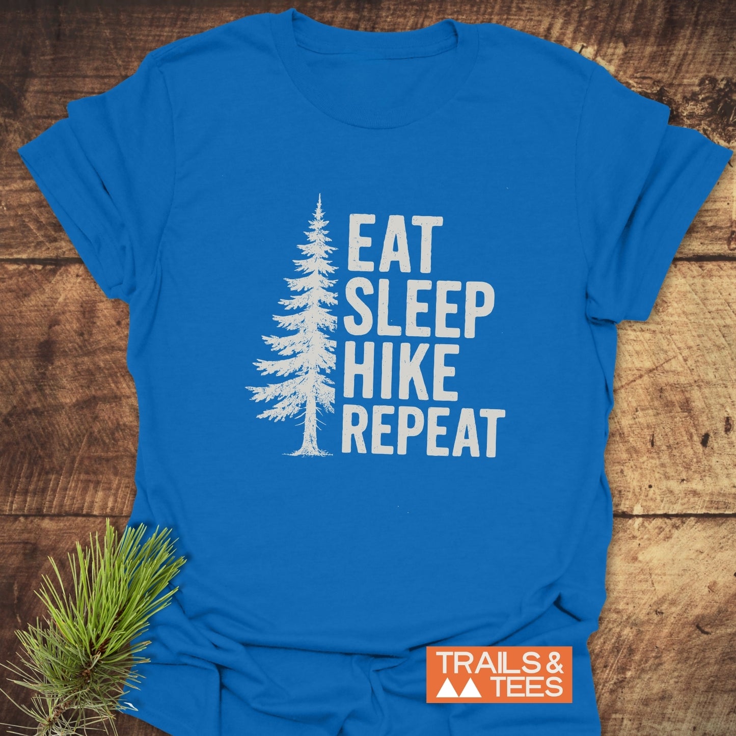 Hike, Repeat T-Shirt with pine tree design and slogan, laid on wood. Features comfort-focused, durable cotton, and Trails & Tees logo. Perfect for outdoor enthusiasts.