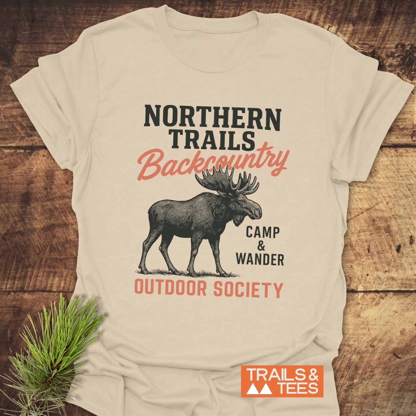 Northern Trails Moose T-Shirt featuring a moose silhouette and bold text, laid on wood. Soft cotton, durable prints, promoting outdoor adventure. Brand: TRAILS & TEES.