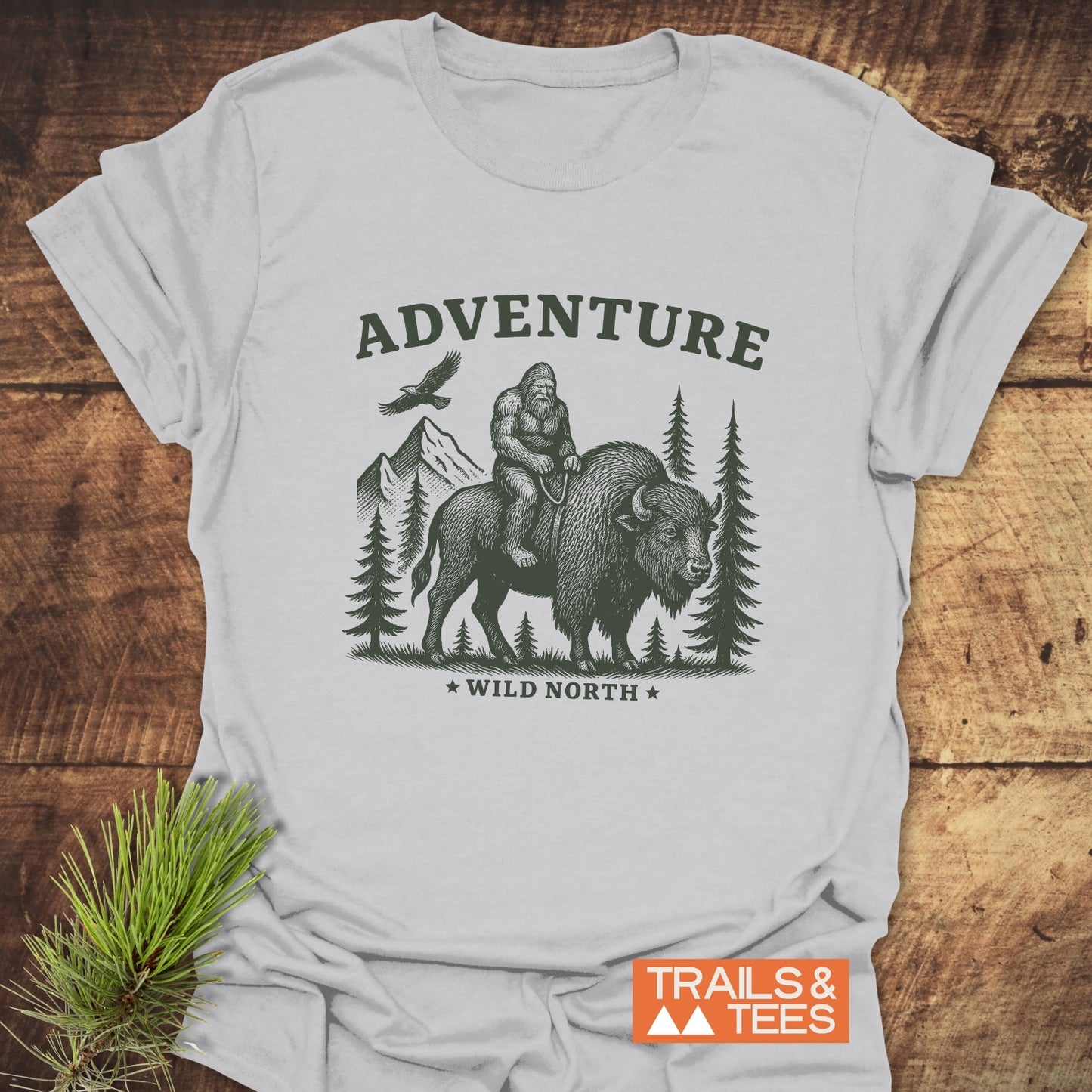Bigfoot & Bison T-Shirt features a woodcut-style Sasquatch atop a bison with mountain scenery, emphasizing adventure. Made from soft, ring-spun cotton for comfort.